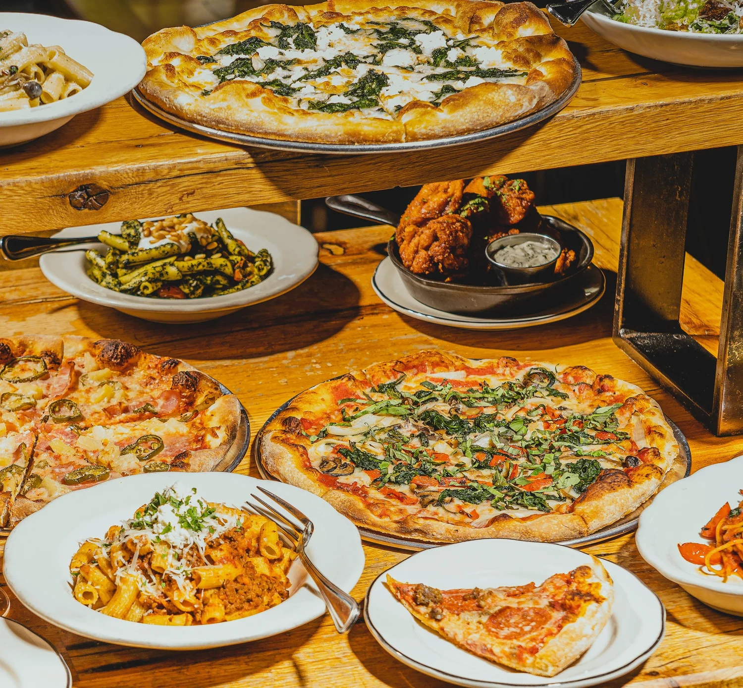 A wooden table crowded with pizzas, pasta, a plate of fried chicken and greens, photographed from above